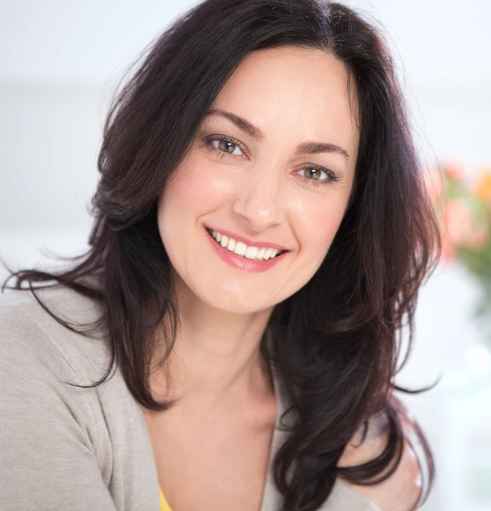 Cosmetic dermatology: A woman with smooth, glowing skin smiling softly while sitting in a bright, cozy environment.